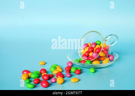 Colored glazed candy beans. Glass mug on saucer and scattered colorful button-shaped chocolates on blue background with copy space. Concept sweet tea Stock Photo