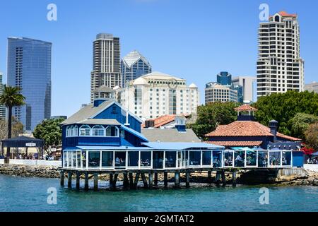 The San Diego Pier restaurant in blue and white retro style on stilts in Seaport Village, with looming high-rise buildings on bay of San Diego, CA Stock Photo