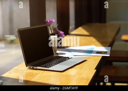 Opened laptop computer black screen mockup and paperworks on wooden table with sunlight through window in coffee shop Stock Photo