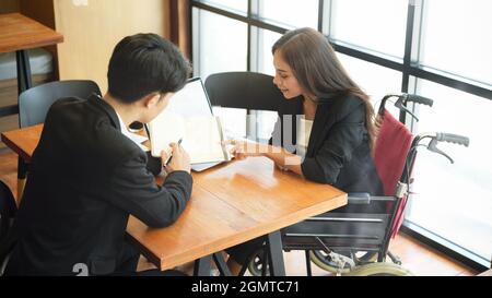 Disabled female business employee in wheelchair brainstorms with coworkers, listen colleague explain work process on notebook in office Stock Photo