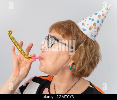 Portrait of a smiling elderly woman in a festive cap holding a whistle tongue on a white background Stock Photo