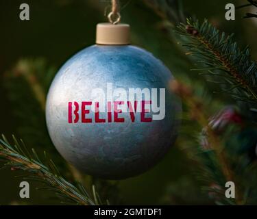 Metal Christmas bulb with BELIEVE written in red letters hanging on a live Christmas tree with blurred background. Stock Photo