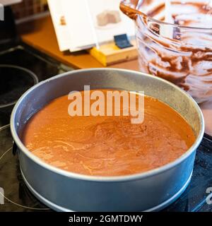 Chocolate cake batter in a metal springform pan with empty glass mixing bowl and cookbook in background out-of-focus. Stock Photo