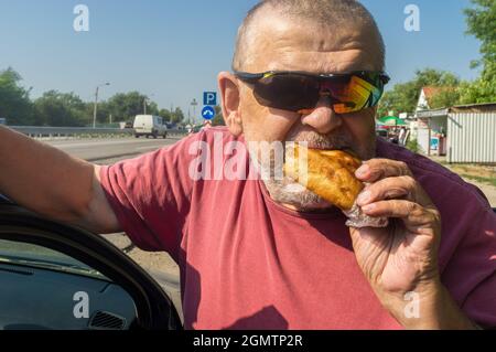 Portarait of hungry Cacasiam senior driver wearing sunglasses eating patty near his car standing on Ukrainian road Stock Photo