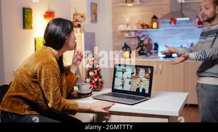 Business woman using video call on laptop working on christmas eve. Adult talking to workmates while man feeling impatient about holiday celebration in festive decorated kitchen. Stock Photo
