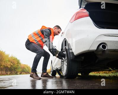 Roadside assistance worker removing flat tire while repairing car on the street. Male auto mechanic in work vest changing flat tire on the road. Concept of emergency road service. Stock Photo