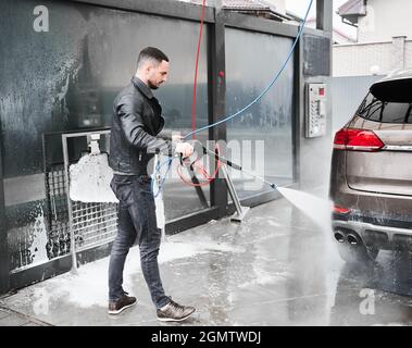 Young man washing car on carwash station outdoor. Handsome worker cleaning automobile, using high pressure water. Stock Photo