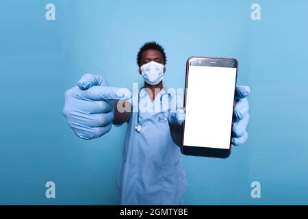 Black nurse pointing at blank white screen on smartphone. African american medical assistant with face mask and stethoscope presenting empty isolated background and mockup template on phone Stock Photo