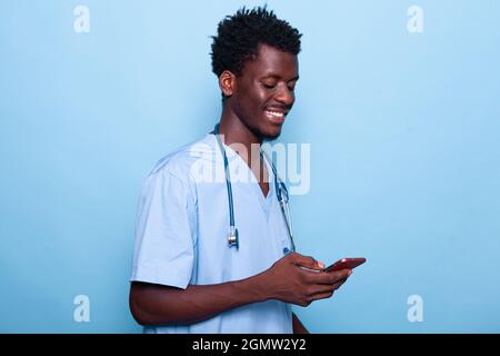 Man working as nurse looking at smartphone and smiling. Medical specialist wearing uniform and stethoscope holding modern phone with touch screen. Healthcare assistant using device Stock Photo