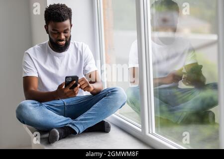 Handsome african man sits home on windowsill, reads text messages, sends email, uses his phone Stock Photo