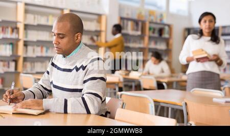 Concentrated adult hispanic man spending time in library, reading books and making notes. Self-education concept Stock Photo