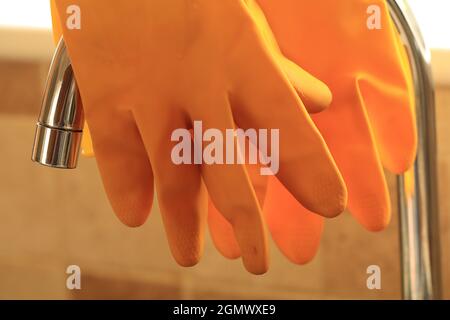 pair of yellow washing up gloves hanging over a tap, tiled background out of focus Stock Photo