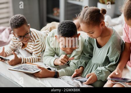 Happy African-American children using gadgets in row while lying on bed together, multiple siblings concept Stock Photo