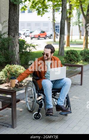 smiling disabled man holding coffee to go while sitting in wheelchair with laptop Stock Photo