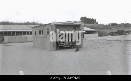 1950s, historical, outside on a wide, level area at a light industrial estate, a man in a suit lifting up the roll-up door of prefabricated garage,   constructed there for demonstration purposes, Witney, Oxford, England, UK. These garages or motor shelters were made from precast concrete panels bolted together into sections, with a corrugated metal roof and metal roll-up or up and over door. As car ownership increased in the 1950s and 60s, these self-build or concrete kit garages became the thing to have and a large number of companies offered such DIY garages to the 'practical householder'. Stock Photo