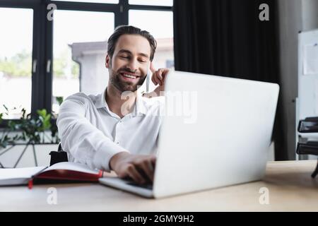 happy businessman using blurred laptop while talking on smartphone in office Stock Photo