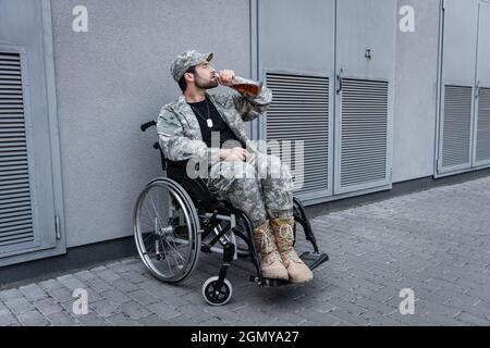 young handicapped veteran sitting in wheelchair on city street and drinking whiskey Stock Photo