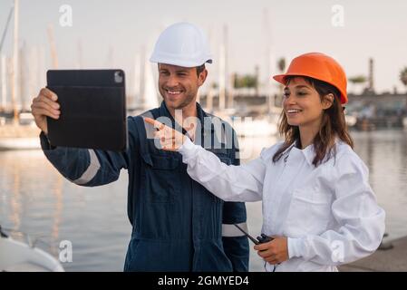 Optimistic young male and female engineers in helmets using tablet and discussing draft while inspecting sea port together Stock Photo