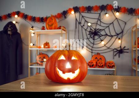 Close up of glowing Halloween pumpkin standing on table in room with other holiday decor. Stock Photo