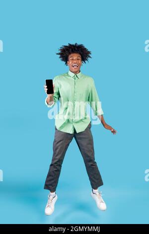Happy black guy showing empty smartphone screen while jumping up over blue studio background, full length Stock Photo