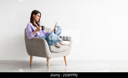 Young Caucasian woman with coffee sitting in armchair and using tablet computer against white studio wall Stock Photo