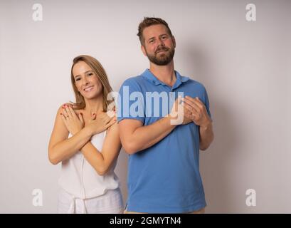 Grateful millennial couple looking at camera holding hands on chest feeling thankful pose isolated on white studio background expressing heartfelt lov Stock Photo