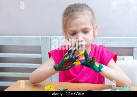 Little girl with colorful painted hands. A concentrated girl smears paints on her hands and looks at the result. The child is learning to paint. Learn Stock Photo