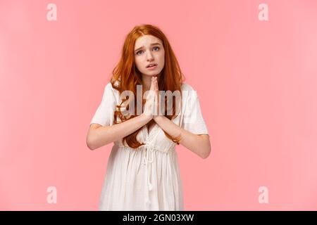 Pleading cute and silly redhead girl acting innocent, press hands together in pray, asking for favour, hopefully looking at camera, frowning gloomy Stock Photo