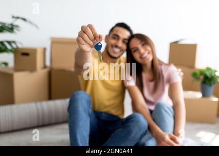 Positive young multiracial couple showing house key, sitting on floor of new home, selective focus Stock Photo