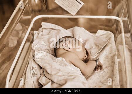 A newborn baby with a maternity hospital bracelet on his arm is sleeping in a crib. A newly born child in a clinic bed behind a transparent glass Stock Photo
