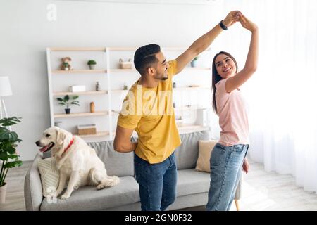 Romantic young diverse couple dancing at home, their cute dog sitting on couch nearby, copy space Stock Photo