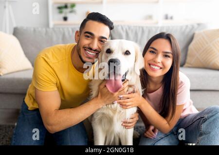 Portrait of smiling diverse couple hugging their pet dog, smiling at camera, indoors Stock Photo
