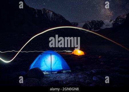 Blue and yellow gloving tent with light line at camping in the mountains under night sky with stars Stock Photo