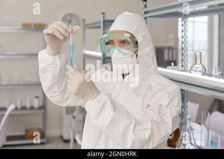 Scientist in coveralls, gloves, face mask and goggles looking at glass tube in his hands Stock Photo