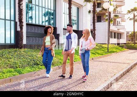 Full body of cheerful young multiracial students in trendy casual outfits chatting and laughing friendly while strolling together on pavement near mod Stock Photo