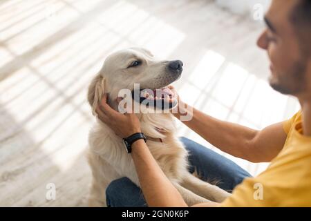 Best friend concept. Millennial Arab man scratching his dog on floor at home, selective focus Stock Photo