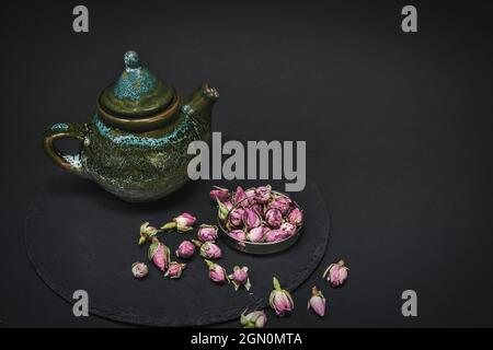 Dried tea rose buds and ceramic vintage teapot for tea ceremony in chinese style on black background. Stock Photo