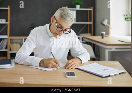 Serious senior businessman sitting at office desk and taking notes in his notebook Stock Photo