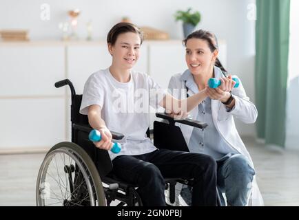 Female physiotherapist assisting happy teen boy in wheelchair to do exercises at home. Handicapped youth training with dumbbells, working out arm musc Stock Photo