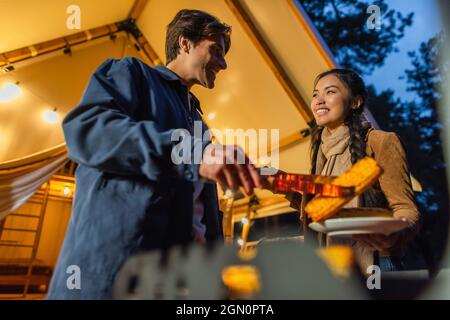 Low angle view of smiling interracial couple cooking on grill near glamping house in evening Stock Photo