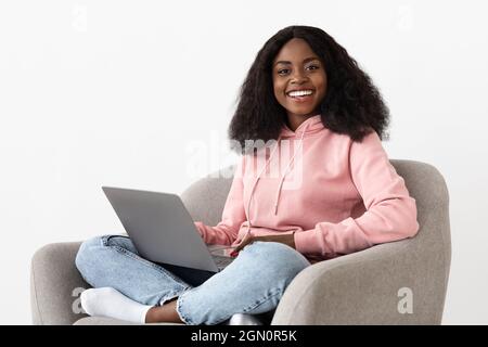 Portrait of millennial black woman independent contractor sitting in stylish arm chair, using laptop, cheerfully smiling at camera, working from home, Stock Photo