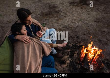 High angle view of smiling interracial couple in blankets looking at campfire Stock Photo