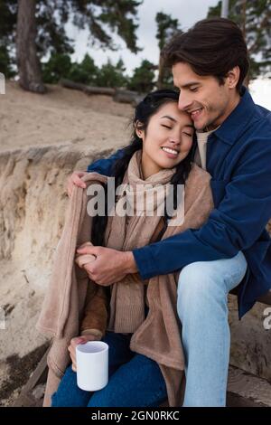 Man wrapping in blanket asian girlfriend with cup near hill Stock Photo