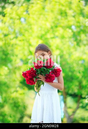 Little cute girl with peony flowers. Child wearing whiye dress playing in a summer garden. Kids gardening. Children play outdoors. Toddler kid with fl Stock Photo