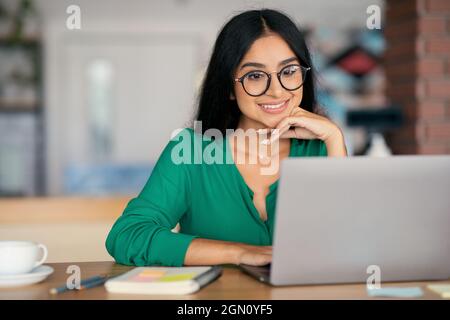 Stylish indian young woman independent contractor wearing glasses working online, looking at laptop screen and smiling, sitting at table, drinking cof Stock Photo