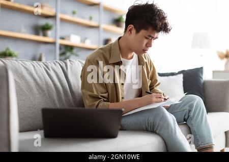 Focused asian man taking notes while working on laptop computer at home office, sitting on sofa. Freelancer noting down information from internet, pla Stock Photo