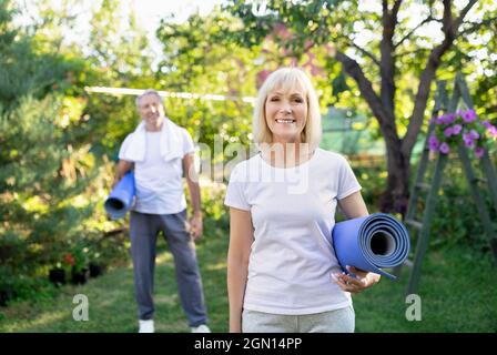 Healthy retirement lifestyle. Happy active senior couple holding yoga mats, looking to camera in sunny garden Stock Photo
