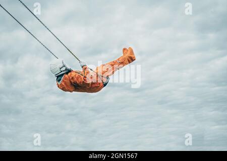 Astronaut wearing orange space suit and space helmet on a swing against cloudy sky Stock Photo