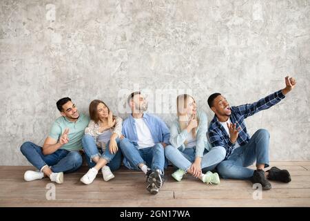 Full length of young diverse students sitting near grey studio wall, taking selfie with cellphone, copy space Stock Photo