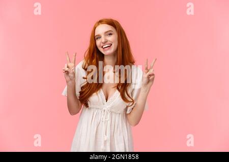 Kawaii cute redhead caucasian girl in white dress showing positivity and joy, laughing smiling carefree, showing peace gestures standing over pink Stock Photo